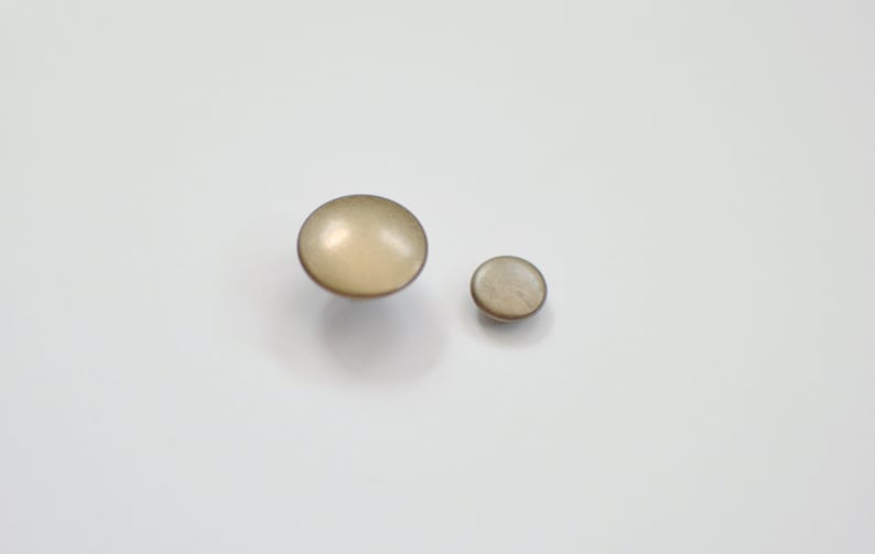 Pu&ograve; includere: Due bottoni rotondi di colore beige con una finitura lucida. Il bottone pi&ugrave; grande &egrave; leggermente pi&ugrave; grande del pi&ugrave; piccolo.