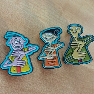 May include: Three cartoon character enamel pins featuring Edd, Eddy, and Double D from the animated television series Ed, Edd n Eddy. The pins are blue, yellow, orange, green, and black.