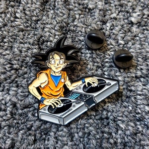 May include: A silver and black enamel pin featuring an anime character with spiky black hair, wearing an orange sleeveless shirt, and a blue undershirt, holding a pair of turntables.