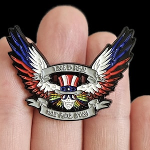 May include: A colorful enamel pin featuring a skull wearing a top hat, with wings in red, white, and blue. The pin has the text "LOVE IS REAL" and "THAT FADE AWAY" on banners. The design is set against a black background.