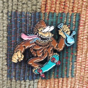 Op de afbeelding: Een emaille pin met een cartoon gorilla in bruin en zwart, die op een blauw-groen skateboard rijdt en een blauwe glazen bong vasthoudt.
