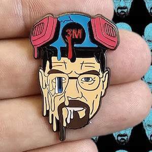 May include: A Breaking Bad Walter White hat pin. The pin features a stylized image of Walter White with a respirator and the text "3M". The pin is primarily black, with accents of red, blue, and yellow.