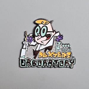 May include: Enamel pin featuring Dexter from Dexter's Laboratory. The pin shows Dexter in his lab coat, holding a beaker. The pin has the words "Dexter's Laboratory" in a stylized font. The pin is on a gray background.