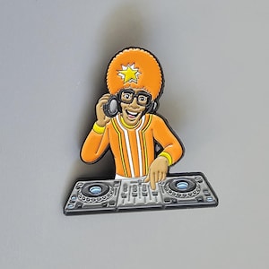 Puede incluir: Un pin de esmalte metálico que muestra a un DJ de dibujos animados con un gran afro naranja, gafas negras y una camisa naranja de manga larga, sosteniendo auriculares en una oreja mientras opera una consola de mezclas gris con dos platos.
