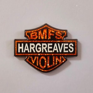 May include: A black and orange pin with the text "BMFS HARGRAVES VIOLIN" in white letters.