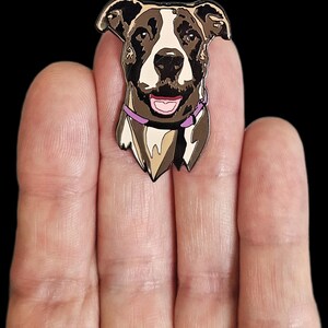 May include: A dog-shaped enamel pin featuring a smiling dog with brown, white, and black markings, wearing a purple collar. The pin is held between fingers against a black background. The dog's tongue is pink.