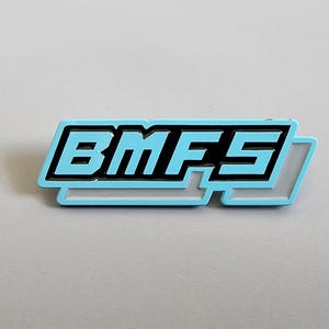 May include: A rectangular pin featuring the letters "BMFS" in black, with a light blue outline, set against a light grey backdrop. The pin has a stylized design with a light blue border.