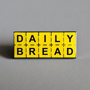 May include: A rectangular pin with the words "DAILY BREAD" in black capital letters on a yellow background. The pin has a black border and is set against a gray backdrop.
