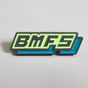 May include: A rectangular pin with the letters "BMF5" in a neon green font, outlined in black. The pin has a gray border and a teal blue accent on the right side. The pin is on a light gray surface.