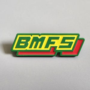 May include: A rectangular pin featuring the letters "BMFS" in yellow, outlined in green, and set against a red background. The pin is on a grey surface.