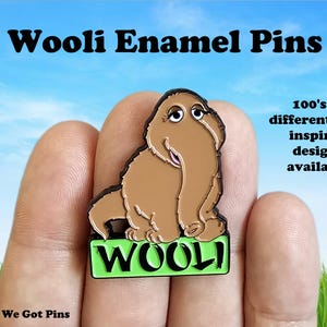 May include: An enamel pin featuring a brown, furry, elephant-like creature with long eyelashes and a trunk, standing above a bright green rectangular base with the black text WOOLI. The text Wooli Enamel Pins and 100's of different EDM inspired designs available appear in the image.