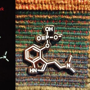 May include: A white glow-in-the-dark enamel pin featuring a chemical structure with the text "O=P-O-" and "OH" on the top and "HN" and "N" on the bottom.