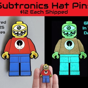 May include: A set of Subtronics Hat Pins featuring a cartoon character design. The pins have a yellow head with a single eye, a red shirt, blue pants, and glow-in-the-dark features. The text "Limited To 25 Pieces" is visible.
