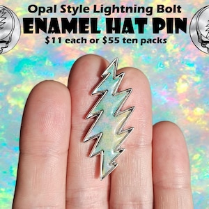 May include: A silver enamel lightning bolt pin with a green, blue, and white opal-like design. The pin is on a finger, and the text "Opal Style Lightning Bolt ENAMEL HAT PIN £9 each or £44 ten packs" is visible in the background.