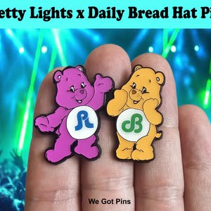 Puede incluir: Dos pines de esmalte para sombrero con personajes de osos de dibujos animados. Un oso es morado con un logotipo azul en su insignia blanca del vientre, y el otro es naranja con un logotipo verde en su insignia blanca del vientre. El texto Pretty Lights x Daily Bread Hat Pins es visible.