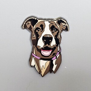 May include: A dog-shaped pin with a brown, white, and black color scheme. The dog has a pink collar and a smiling expression. The pin is likely made of metal or a similar material and is designed to be attached to clothing or accessories.