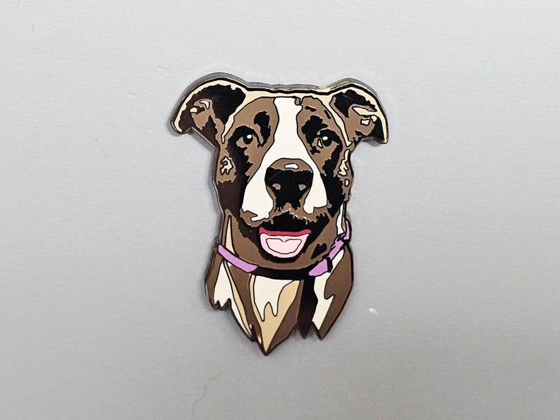 May include: A dog-shaped pin with a brown, white, and black color scheme. The dog has a pink collar and a smiling expression. The pin is likely made of metal or a similar material and is designed to be attached to clothing or accessories.