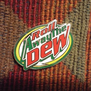 May include: A green, yellow and red enamel pin with the words "Roll Away the Dew" in a white oval.