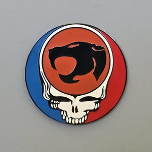 May include: A round, colorful pin featuring a stylized skull with a red and blue border. Inside the red circle is a black panther silhouette. The pin is a vibrant accessory.