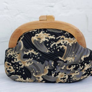 Puede incluir: Un bolso de mano con marco de madera y tela negra con un estampado de olas en tonos grises y dorados. El cierre es de madera clara. El bolso está diseñado para llevarse en la mano.
