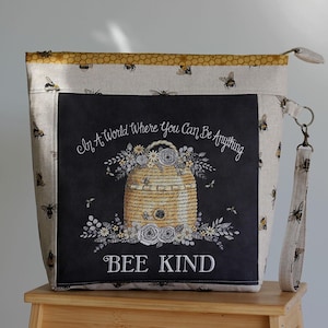 Könnte beinhalten: Eine Reißverschlusstasche aus beigem Stoff mit Bienenmuster und gelbem Wabenrand. Das schwarze Mittelteil zeigt eine Grafik eines Bienenstocks aus Stroh, umgeben von Blumen und dem Text In A World Where You Can Be Anything BEE KIND.