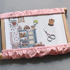 Könnte beinhalten: Ein Kreuzstichprojekt in einem Holzrahmen mit einem rosa Stoffrand. Der Kreuzstich zeigt einen blauen Schrank mit Teddybären und anderen Gegenständen. Eine Schere und Holzverzierungen befinden sich auf dem weißen Stoff.