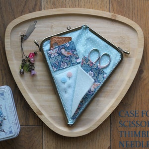 Könnte beinhalten: Ein hellblaues, dreieckiges Nähset mit Blumenmuster, das eine Schere, Nadeln und einen Fingerhut enthält. Das Set liegt auf einem Holztablett, neben einer dekorativen Dose. Ein Anhänger mit Perlen hängt daran.