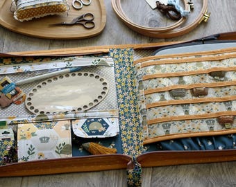 Puede incluir: Kit de costura abierto en amarillo mostaza con motivos florales, con compartimentos para agujas, hilos y un marco ovalado de madera. Incluye tijeras, bandeja de madera y aros de bordado. El interior es azul. Varios accesorios de costura son visibles.