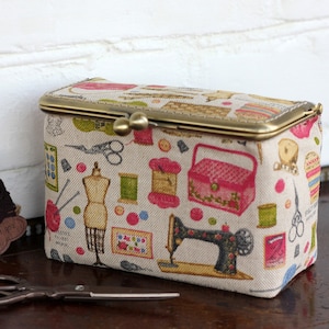May include: A rectangular fabric storage box with a brass kiss-lock closure, featuring a beige background printed with colorful sewing-themed illustrations including a sewing machine, dress form, scissors, spools of thread, and a pink sewing box.