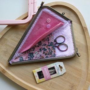 May include: A pink fabric triangle sewing kit with a zip closure. The kit is open and contains a pair of small scissors, a small pack of needles, and a pink felt piece with two buttons. The kit is sitting on a wooden surface.