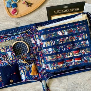 Op de afbeelding: Een marineblauwe stoffen opbergmap met een herhalend Alice in Wonderland-patroon en witte stippenrand. De binnenkant bevat meerdere vakjes, elastische lussen, een kleine houten borduurring, een schaar en een gouden kwastje aan de rits.