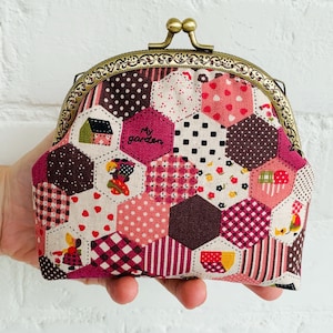 Puede incluir: Un monedero de patchwork con cierre metálico. El monedero presenta una variedad de hexágonos de tela estampada en tonos rosa, burdeos y blanco. La frase "My garden" es visible en la parte delantera. El cierre es de color bronce decorativo.
