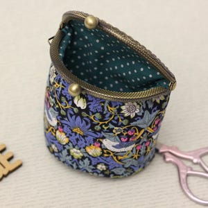 May include: A fabric pouch with a kiss-lock bronze metal frame, featuring a dark blue floral and bird pattern on the exterior and a teal interior lining with white polka dots. The pouch is open, revealing the interior fabric and metal clasp.