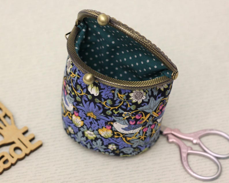 May include: A fabric pouch with a kiss-lock bronze metal frame, featuring a dark blue floral and bird pattern on the exterior and a teal interior lining with white polka dots. The pouch is open, revealing the interior fabric and metal clasp.
