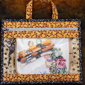 Puede incluir: Una bolsa de proyecto transparente con cremallera, con un patr&oacute;n de panal y un borde negro. La bolsa contiene un proyecto de punto de cruz, hilo de bordar y unas tijeras.