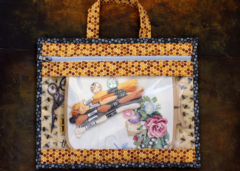Puede incluir: Una bolsa de proyecto transparente con cremallera, con un patr&oacute;n de panal y un borde negro. La bolsa contiene un proyecto de punto de cruz, hilo de bordar y unas tijeras.