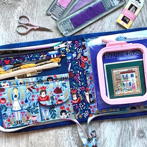 Pode incluir: Um organizador de artesanato de tecido azul com zíper, cheio de materiais de bordado. Contém carretéis de linha, um bastidor de bordado rosa, tesouras e um projeto de ponto cruz. O organizador tem bolsos com tecido temático Alice no País das Maravilhas.
