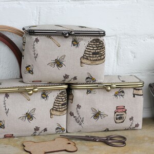 May include: Three rectangular fabric pouches with metal kiss-lock frames, featuring a beige background with a repeating pattern of bees, beehives, honey jars, and floral sprigs. The pouches have metallic closures in dark and brass finishes.