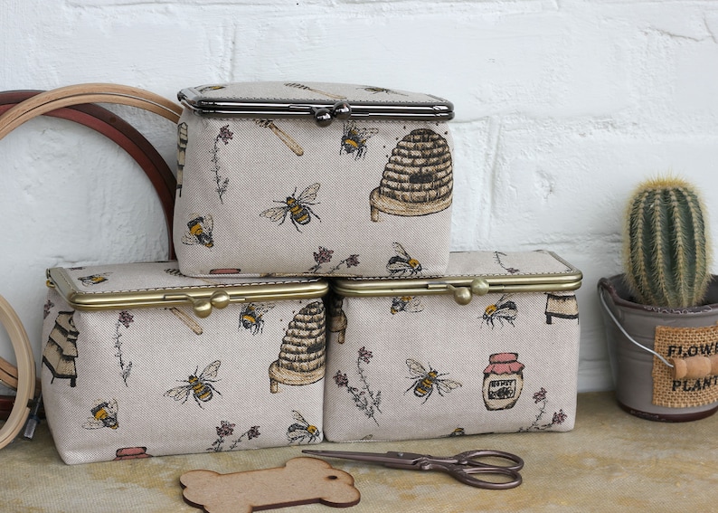 May include: Three rectangular fabric pouches with metal kiss-lock frames, featuring a beige background with a repeating pattern of bees, beehives, honey jars, and floral sprigs. The pouches have metallic closures in dark and brass finishes.