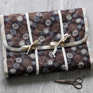May include: A quilted, brown fabric case with a floral pattern in shades of brown, white, and blue. The case is secured with cream-colored ribbons and wooden buttons. A pair of small, silver scissors rests nearby.