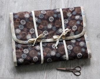 May include: A quilted, brown fabric case with a floral pattern in shades of brown, white, and blue. The case is secured with cream-colored ribbons and wooden buttons. A pair of small, silver scissors rests nearby.