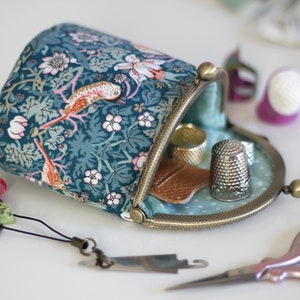 May include: A fabric pouch with a teal floral and bird pattern, featuring a bronze-toned metal kiss-lock frame. The open pouch contains a silver thimble and a small brown leather piece, with sewing accessories nearby.