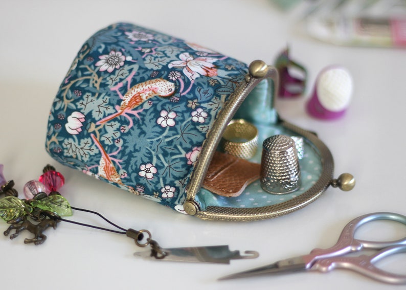 May include: A fabric pouch with a teal floral and bird pattern, featuring a bronze-toned metal kiss-lock frame. The open pouch contains a silver thimble and a small brown leather piece, with sewing accessories nearby.