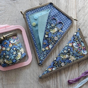 May include: A pink embroidery hoop holds a small, round, blue floral fabric container with a metal clasp. Two triangular fabric cases and a third container with the same floral pattern are also present. The cases have metal clasps and hold scissors and felt.