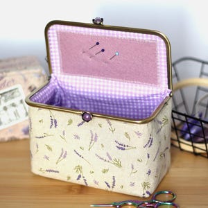 May include: A rectangular sewing box with a lavender floral pattern on a cream background. The box has a gold-colored frame and a purple interior with a gingham pattern. The lid is open, revealing pins. A pair of scissors is in the foreground.