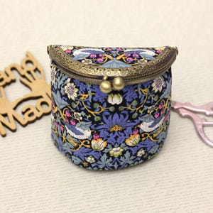 Op de afbeelding: Een kleine, ronde stoffen tas met een bloemen- en vogelpatroon in blauw, wit en geel. De tas heeft een bronskleurige sluiting en twee goudkleurige bollen. Een houten uitsnede met de tekst "Hand Made" staat links van de tas.