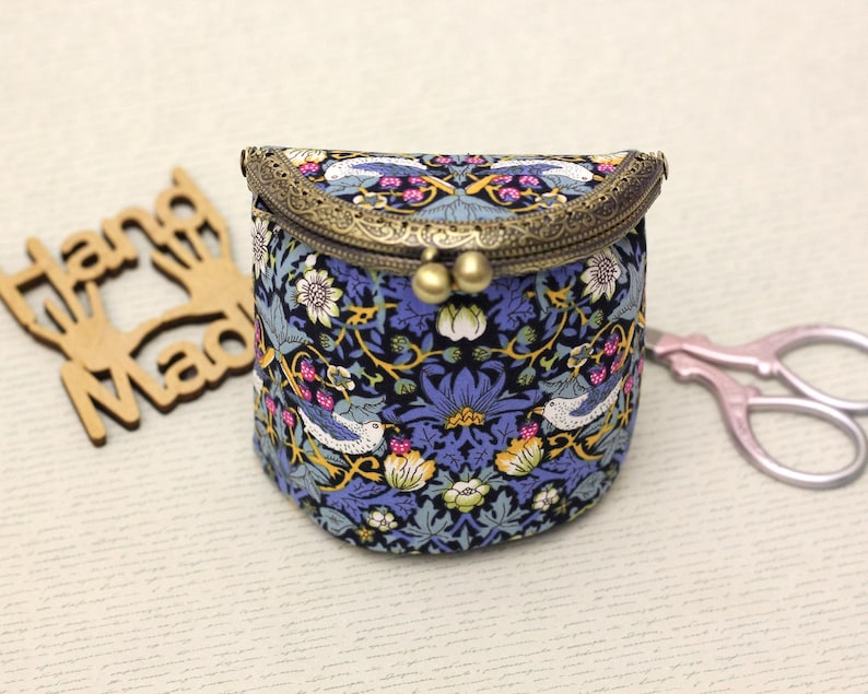 May include: A small, round fabric purse with a floral and bird pattern in blue, white, and yellow. The purse has a bronze-colored clasp and two gold-colored spheres. A wooden cutout that says "Hand Made" is to the left of the purse.