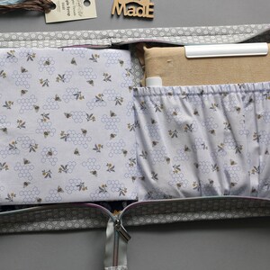 May include: Open grey fabric zippered pouch with a honeycomb and bee pattern lining. The pouch is filled with fabric scraps and a white plastic ruler. The pouch is labeled "Made".