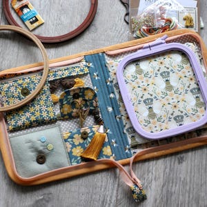 May include: A fabric embroidery organizer case featuring a bee and floral pattern, containing a light purple square embroidery hoop, a felt needle cushion with pins, small scissors, a gold tassel, and zippered pockets for supplies.