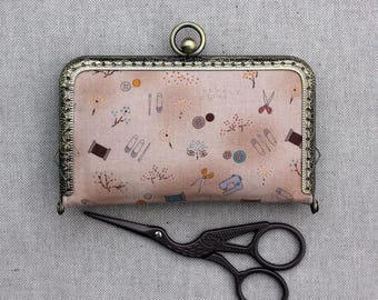 Pode incluir: Uma pequena carteira retangular com fecho de metal e um padrão floral decorativo. A carteira é rosa claro com imagens de artigos de costura. Uma tesoura de bordar preta está por baixo.